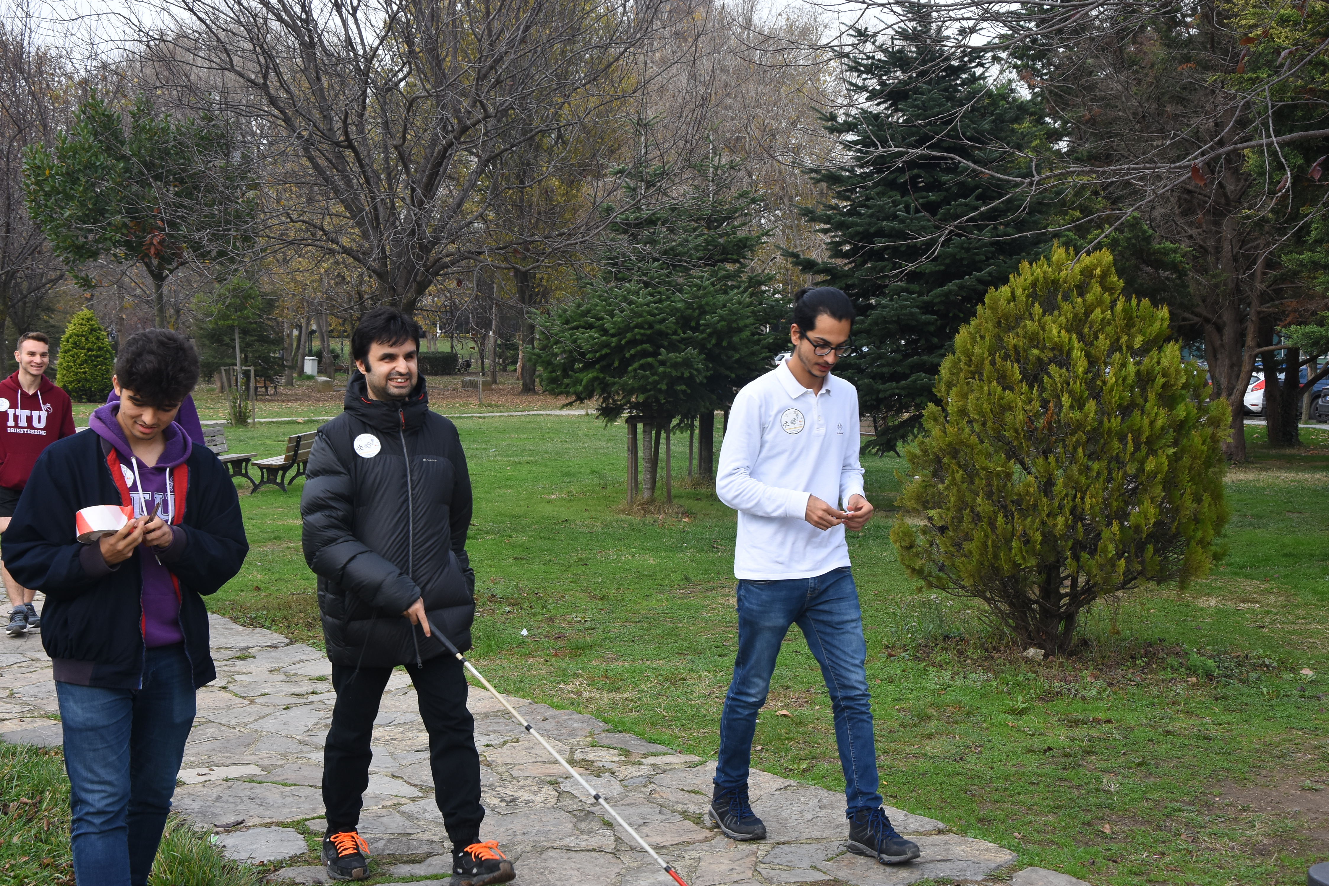 Engelli Öğrenci Birim Koordinatörü Çağrı Artan ve kulüp öğrencilerinin yeşil alan üzerindeki taşlı yolda yürüdükleri, ellerindeki hedef kartları ile parkur hedefine ilerledikleri bir an.